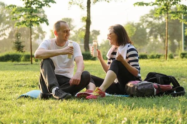 Feliz pareja madura sentada en el parque en la colchoneta de fitness, descansando bebiendo yogur después de ejercicios deportivos . —  Fotos de Stock