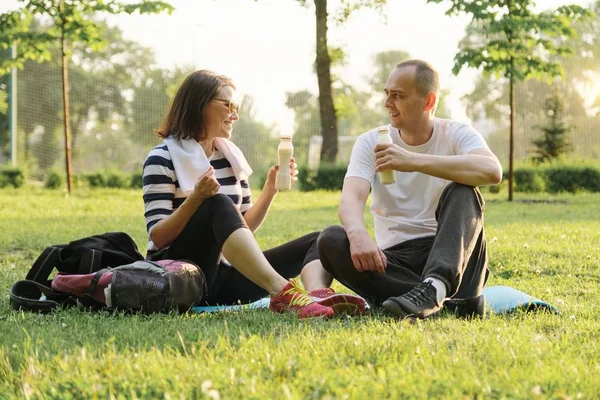 Feliz pareja madura sentada en el parque en la colchoneta de fitness, descansando bebiendo yogur después de ejercicios deportivos . —  Fotos de Stock