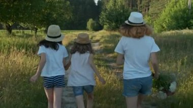 Şapkalı üç kız, kırsal yol boyunca koşarak el ele tutuşuyor