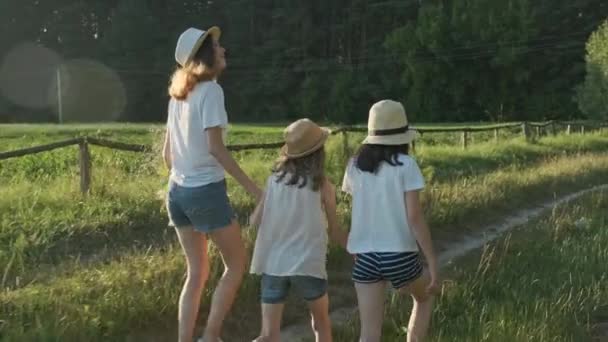 Παιδιά τρία κορίτσια με καπέλα κρατώντας τα χέρια τρέχουν πίσω κατά μήκος του αγροτικού δρόμου — Αρχείο Βίντεο