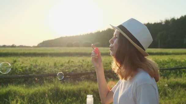 Hermosa chica adolescente en un sombrero soplando burbujas de jabón — Vídeo de stock