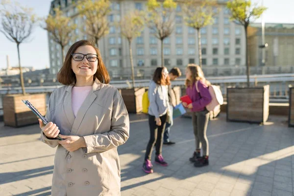 Портрет взрослой улыбающейся учительницы в очках с планшетом, на открытом воздухе с группой подростков — стоковое фото