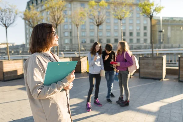 Portrett av en moden, smilende kvinnelig lærer i briller med skriveplate, utendørs med en gruppe tenåringselever – stockfoto