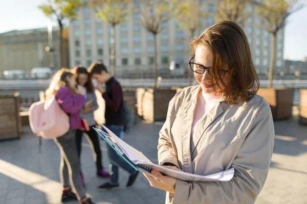 Portrait of mature smiling female teacher in glasses with clipboard, outdoor with a group of teenagers students — Stock Photo, Image