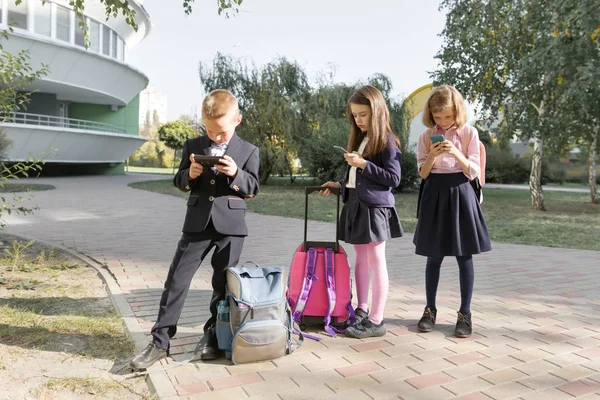 Группа детей 7, 8 лет с мобильными телефонами, школьники с рюкзаками, заглядывающие в смартфоны — стоковое фото