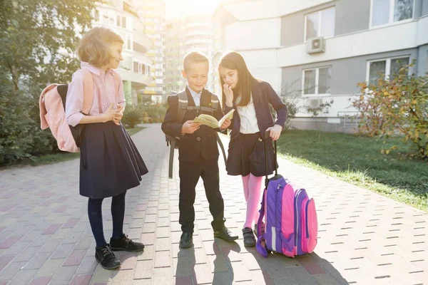 Buiten portret van lachende schoolkinderen op de basisschool. Groep kinderen met rugzakken hebben plezier, praten, het lezen van een boek — Stockfoto