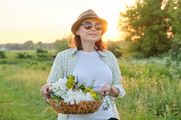 Mogen kvinna som går med en korg och blommor i en sommaräng, gyllene timmen — Stockfoto