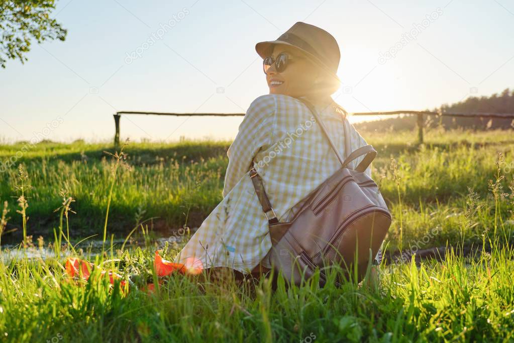 Adult woman in hat with backpack sitting back, summer landscape background