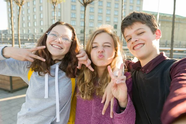 友人の十代の少年と2人の女の子の肖像画は、通りで勝利の兆候を示し、面白い顔を作り、笑顔. — ストック写真