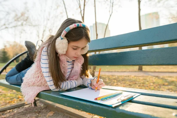 Desenho bonito pequeno do artista com lápis coloridos, menina que senta-se em um banco no parque ensolarado do outono — Fotografia de Stock