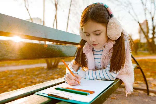 Desenho bonito pequeno do artista com lápis coloridos, menina que senta-se em um banco no parque ensolarado do outono — Fotografia de Stock