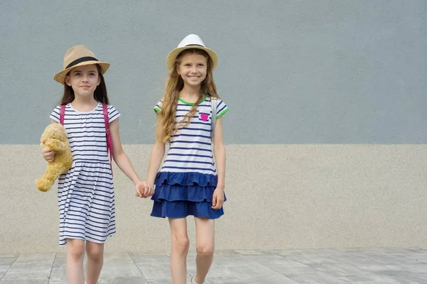 แฟนสาวที่สวยงามสองคนจับมือกัน เด็กผู้หญิงเดินในชุดรัดรูป หมวกพร้อมกระเป๋าเป้ — ภาพถ่ายสต็อก