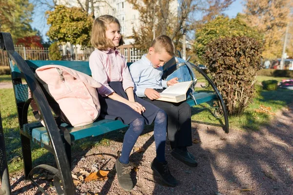 Kleine jongen en meisje schoolkinderen lezen boek, zittend op Bench, kinderen met rugzakken — Stockfoto