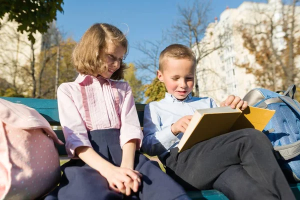 Little boy and girl schoolchildren reading book, sitting on bench, children with backpacks — Stock Photo, Image