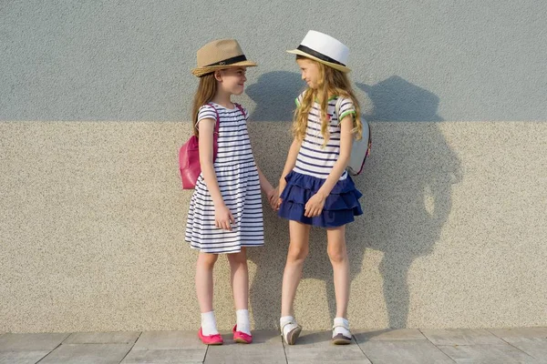 Plener letni portret dwóch szczęśliwych przyjaciółek 7, 8 lat w profilu mówienia i śmiechu. Dziewczyny w pasiastej sukienki, czapki z plecakiem — Zdjęcie stockowe