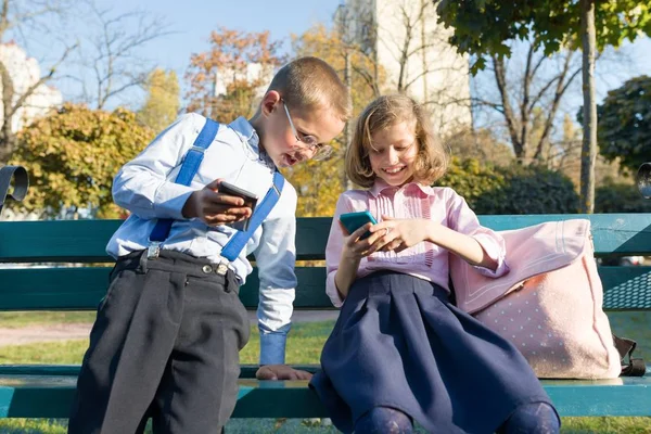 Niños divertidos niño y niña están buscando en los teléfonos inteligentes. En un banco con mochilas escolares, fondo otoño soleado parque —  Fotos de Stock