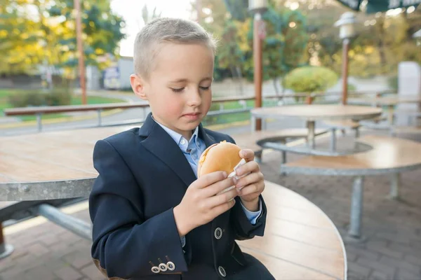 Jongen basisschool student eet hamburger, sandwich in een openluchtcafé. — Stockfoto