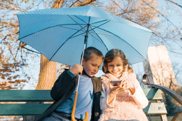 Retrato al aire libre de dos niños sonrientes de niño y niña, sentados bajo un paraguas en el banco del parque, mirando el teléfono inteligente, hora dorada — Foto de Stock