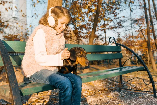 Petit propriétaire souriant d'un chien teckel assis sur un banc dans le parc d'automne, fille avec amour étreignant chien à l'heure d'or — Photo