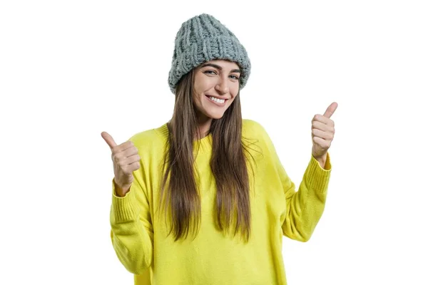 Jovem mulher bonita em suéter amarelo e cinza grande loop tricotado chapéu gorro, mostrando sinal ok polegares para cima, olhando para a câmera no fundo isolado branco — Fotografia de Stock