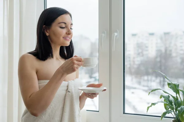 Утренний портрет молодой красивой женщины в банном полотенце с чашкой свежего кофе у окна — стоковое фото