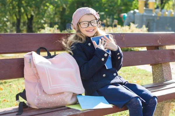 Μικρό κορίτσι μαθητής Δημοτικού σχολείου κάθεται σε παγκάκι με σακίδιο, μοιάζει σε σχολικά βιβλία και τετράδια. Φόντο φθινοπωρινές πάρκο πόλης — Φωτογραφία Αρχείου