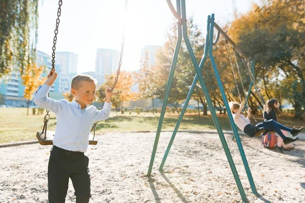 Los niños montan en un columpio en el parque de otoño. Concéntrate en el chico, las chicas en la distancia, hora dorada — Foto de Stock