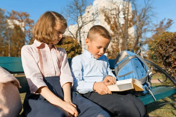 Мальчик и девочка школьники читают книгу, сидят на скамейке, дети с рюкзаками, яркий солнечный осенний день — стоковое фото