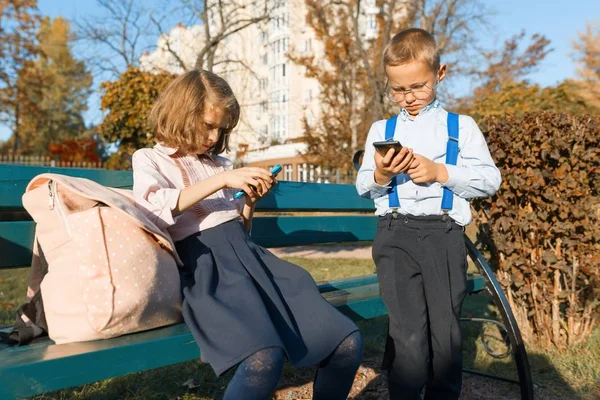 Niños serios inteligentes niño y niña están buscando en los teléfonos inteligentes. En un banco con mochilas escolares, fondo otoño soleado parque, hora dorada —  Fotos de Stock