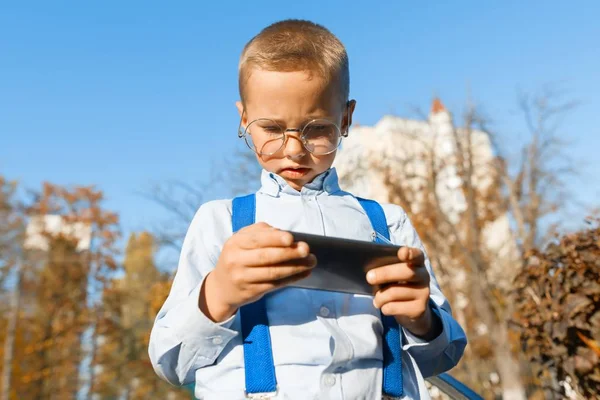 Niño inteligente en gafas con un teléfono móvil. Un niño en una camisa clásica en tirantes para pantalones, fondo del parque soleado, la hora dorada —  Fotos de Stock