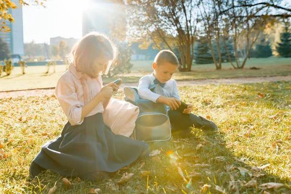 Los niños y niñas de la escuela se sientan en el parque de otoño en la hierba con mochilas y miran los teléfonos inteligentes. Fondo otoño soleado parque, hora dorada — Foto de Stock