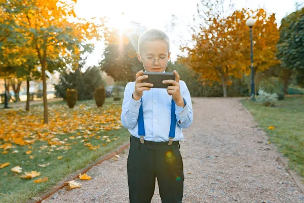 Умный мальчик в очках с мобильным телефоном. Ребенок в классической рубашке в подтяжках для брюк, на фоне солнечного парка, золотой час — стоковое фото