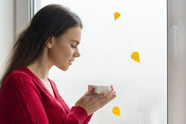 Otoño en la ventana, una joven se para cerca de la ventana con gotas de lluvia y una hoja amarilla, bebiendo café — Foto de Stock