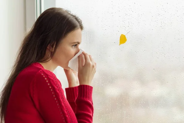 Young woman with handkerchief sneezes, stands near a rainy autumn window, cold season, copy space