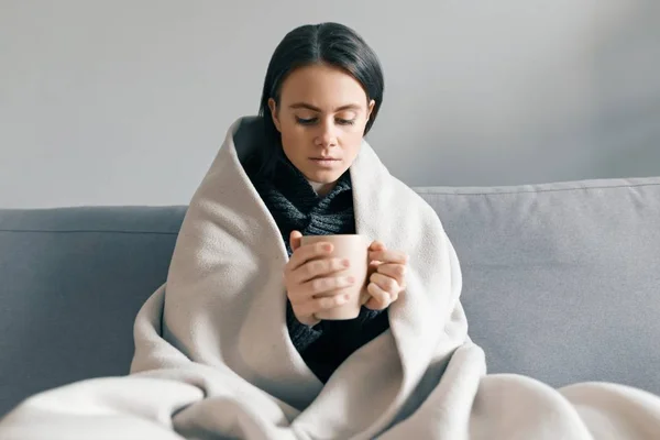 Retrato de invierno de otoño de una joven que descansa en casa en el sofá con teléfono móvil y una taza de bebida caliente, bajo una manta caliente —  Fotos de Stock