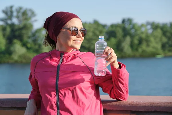 Healthy lifestyle of mature woman, outdoor portrait of an age female in sportswear with yoga mat, drinking water from bottle