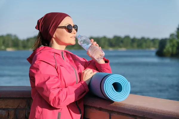 Hälsosam livsstil av mogen kvinna, Utomhus porträtt av en ålder kvinna i sportkläder med yogamatta, dricksvatten från flaska — Stockfoto