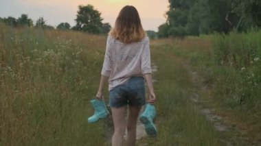 Genç kız elinde botlar ile kırsal yolda yürüyüş. Kirli kıyafetlerle mutlu kız, su birikintisi, arka görünüm, yaz doğası arka planda oyun bulanık