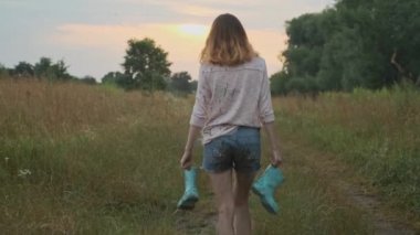 Genç kız elinde botlar ile kırsal yolda yürüyüş. Kirli kıyafetlerle mutlu kız, su birikintisi, arka görünüm, yaz doğası arka planda oyun bulanık