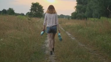 Genç kız elinde botlar ile kırsal yolda yürüyüş. Kirli kıyafetlerle mutlu kız, su birikintisi, arka görünüm, yaz doğası arka planda oyun bulanık