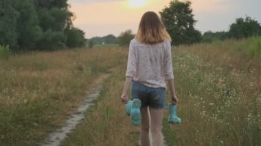 Genç kız elinde botlar ile kırsal yolda yürüyüş. Kirli kıyafetlerle mutlu kız, su birikintisi, arka görünüm, yaz doğası arka planda oyun bulanık