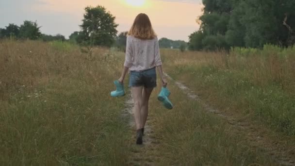 彼女の手にブーツを持って田舎道を歩く十代の少女 汚れた服を着た幸せな女の子 水たまりで遊ぶぼやけた バックビュー 夏の自然の背景 — ストック動画