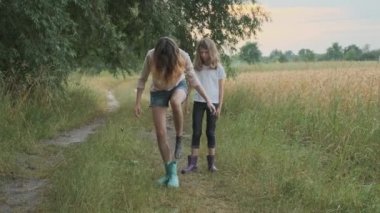 Kirli giysiler içinde yağmurdan sonra yürüyen iki mutlu kız kardeş botlarından su dökerken eğleniyor