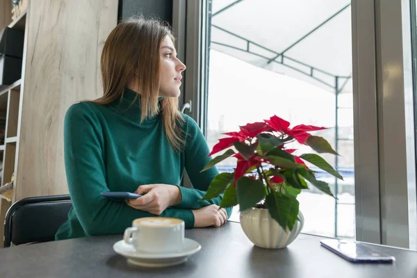 Retrato de invierno de la joven hermosa niña sentada en la cafetería con una taza de café y teléfono móvil, nieve fuera de la ventana, flor roja de poinsettia de Navidad en la mesa — Foto de Stock