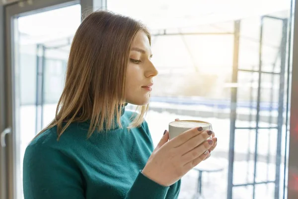 Menina bonita nova bebendo café, de pé no perfil, olhando pela janela. Temporada de inverno de outono no café — Fotografia de Stock