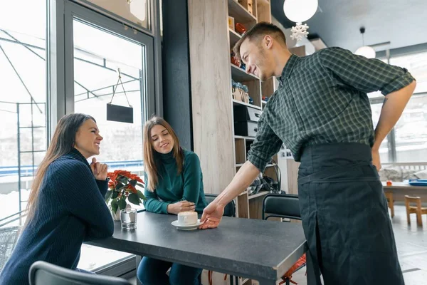 Café, saison d'hiver, deux jeunes femmes assises à la table, et un barista masculin avec une tasse de café souriant et parlant — Photo