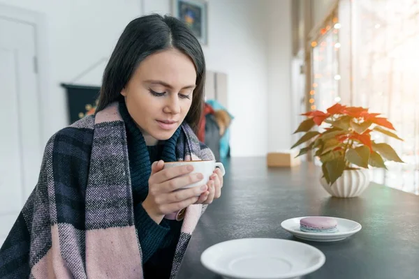 Ung vacker kvinna under vintersäsongen värmer upp med varmt kaffe och varm filt sitter i kafé nära fönstret — Stockfoto