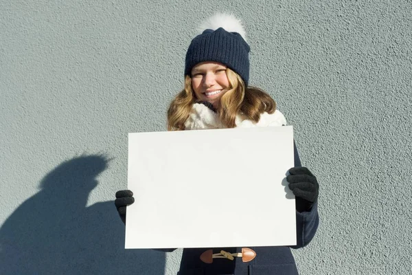 Joven chica sonriente en invierno sostiene una hoja de papel blanco limpio —  Fotos de Stock