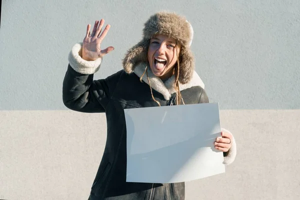 Mladá zábavná dívka v zimních šatech, klobouk zobrazující čistý bílý list papíru — Stock fotografie