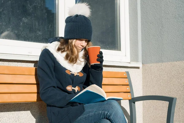 Ritratto invernale di giovane studentessa adolescente in cappello, vestiti caldi con tazza di bevanda calda e libro — Foto Stock